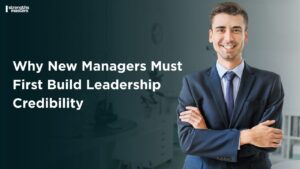 A confident new manager in a formal suit stands with folded arms in an office setting beside the text “Why New Managers Must First Build Leadership Credibility.”